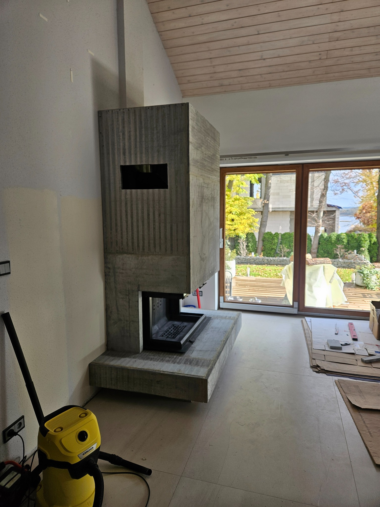 Surowy, betonowy kominek o prążkowanej fakturze w nowoczesnym wnętrzu z dużym oknem i drewnianym sufitem. Żółty odkurzacz Karcher przy ścianie.