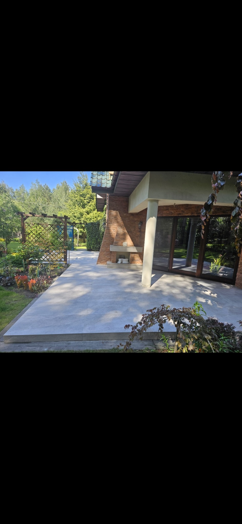 Jasnoszara, gładka posadzka tarasowa z dużych płyt, przylegająca do domu z ceglaną elewacją i ogrodem. Widoczna pergola i roślinność.
