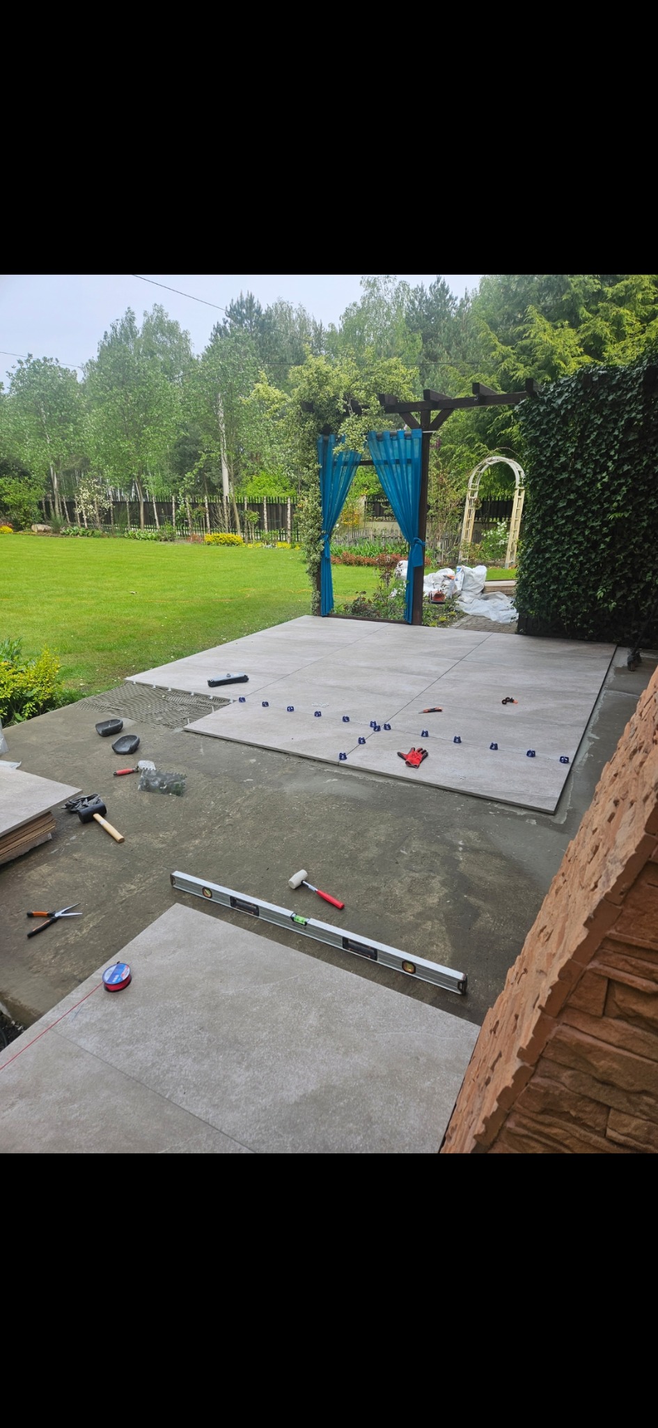 Układanie jasnoszarych płytek na tarasie w ogrodzie. Widoczne narzędzia glazurnicze, poziomica, młotek, sznurek traserski i niebieskie krzyżyki dystansowe.