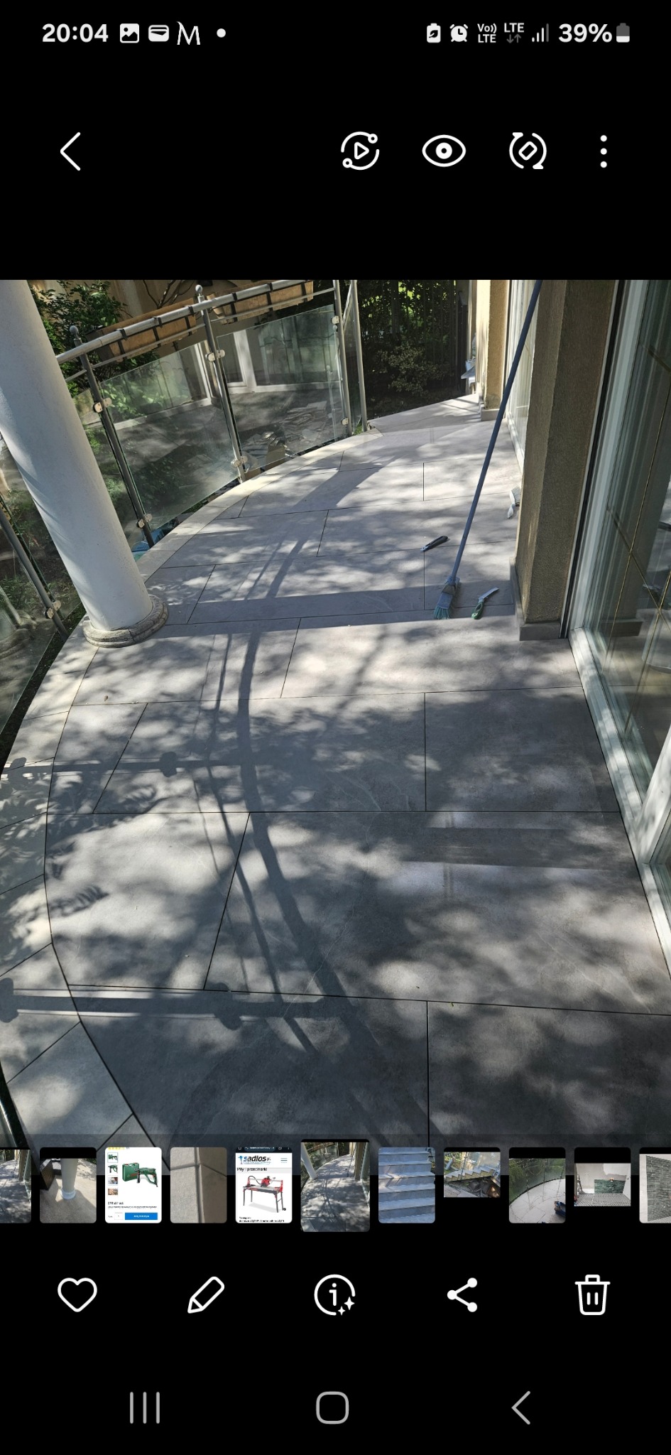 Wyłożony szarymi płytkami taras z szklaną balustradą i kolumną. Na płytkach widoczne cienie drzew. Obok ściany oparta miotła. Widok na zewnątrz.