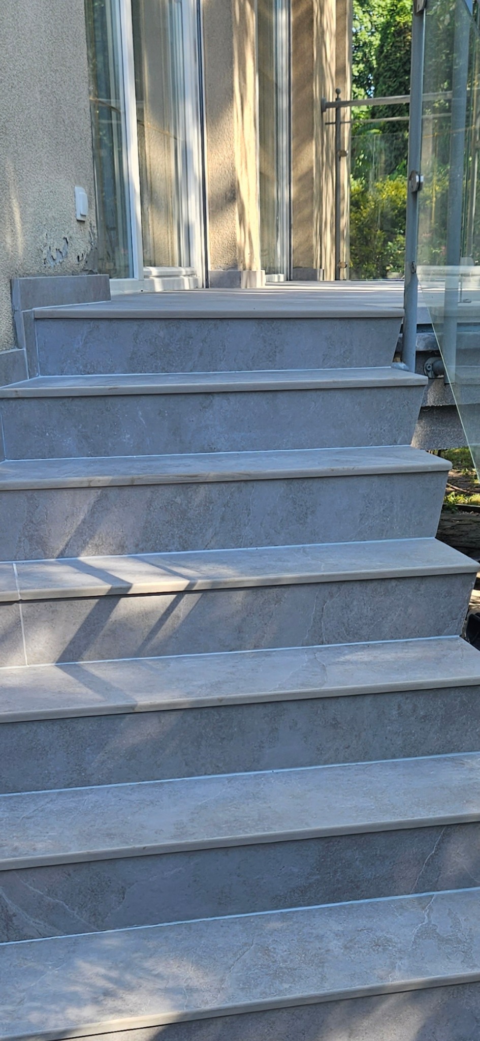 Zewnętrzne schody wyłożone szarymi płytkami z jasnymi krawędziami, prowadzące do przeszklonego wejścia. Widoczne cienie drzew i fragment szklanej balustrady.