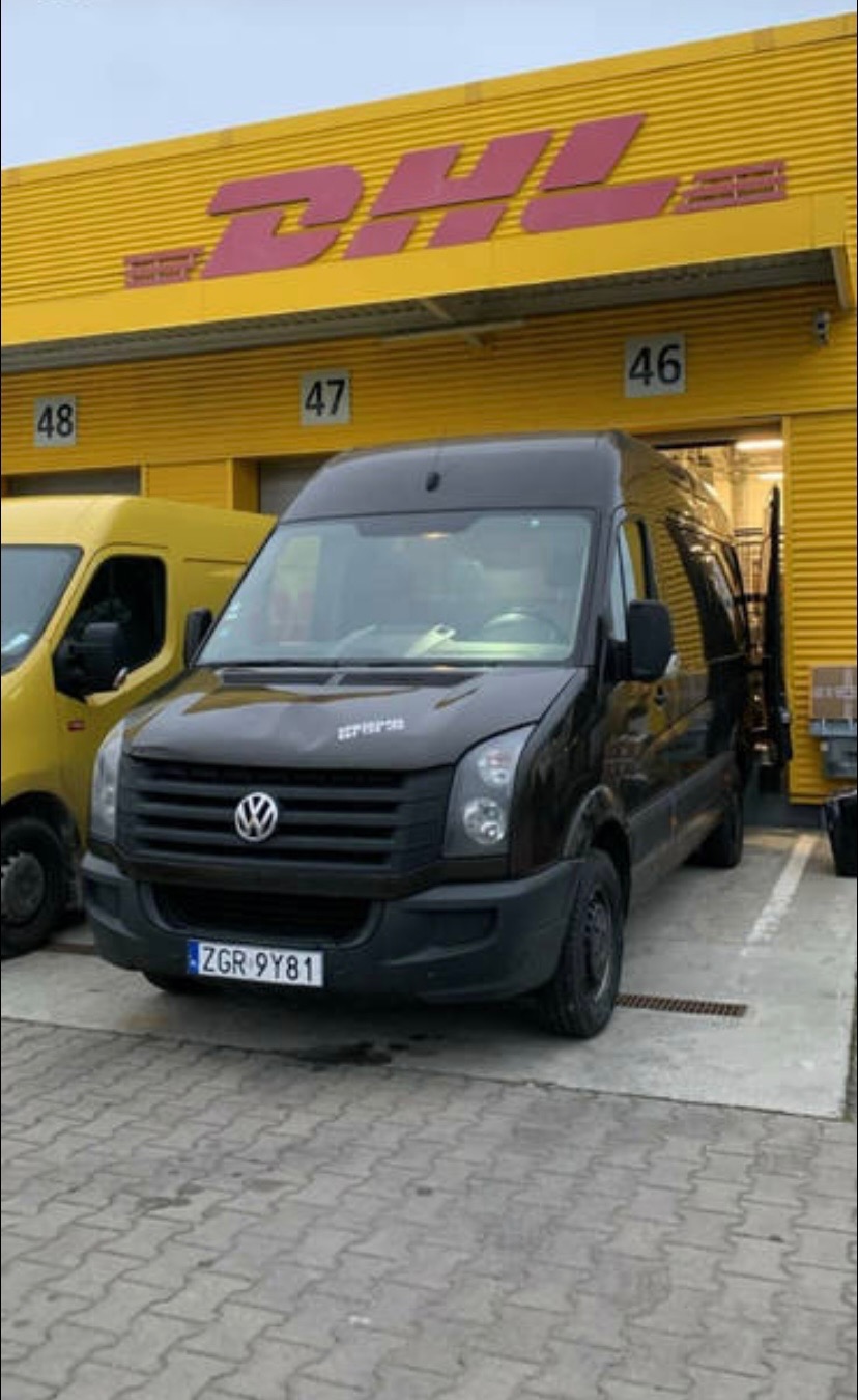 Czarny bus Volkswagen Crafter z numerem rejestracyjnym ZGR 9Y81 zaparkowany przed żółtym budynkiem DHL z numerami 46, 47 i 48 nad wjazdami.
