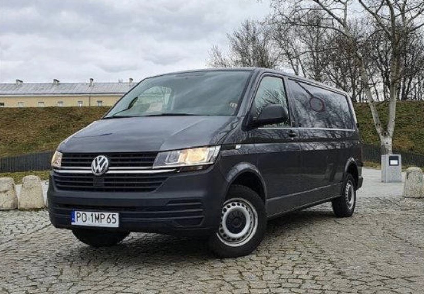 Szary Volkswagen Transporter T6.1 z polską tablicą rejestracyjną PO 1MP65, zaparkowany na brukowanej nawierzchni przed budynkiem z cegły i drzewami bez liści.