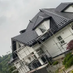 Dom jednorodzinny z czarnym dachem krytym dachówką, białą elewacją i rusztowaniami w trakcie prac wykończeniowych, na tle zielonego wzgórza.