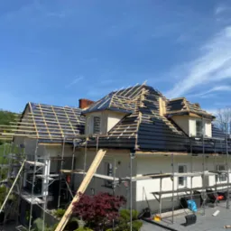 Dom jednorodzinny w trakcie układania czarnej dachówki na drewnianej konstrukcji, widoczne rusztowania i błękitne niebo.