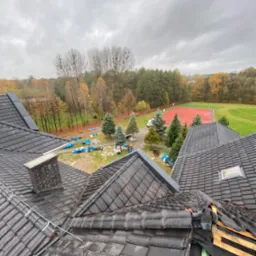 Widok z góry na dach pokryty ciemną dachówką, z fragmentem dachu w trakcie naprawy i widocznym kominem; w tle drzewa w jesiennych barwach i boisko sportowe.