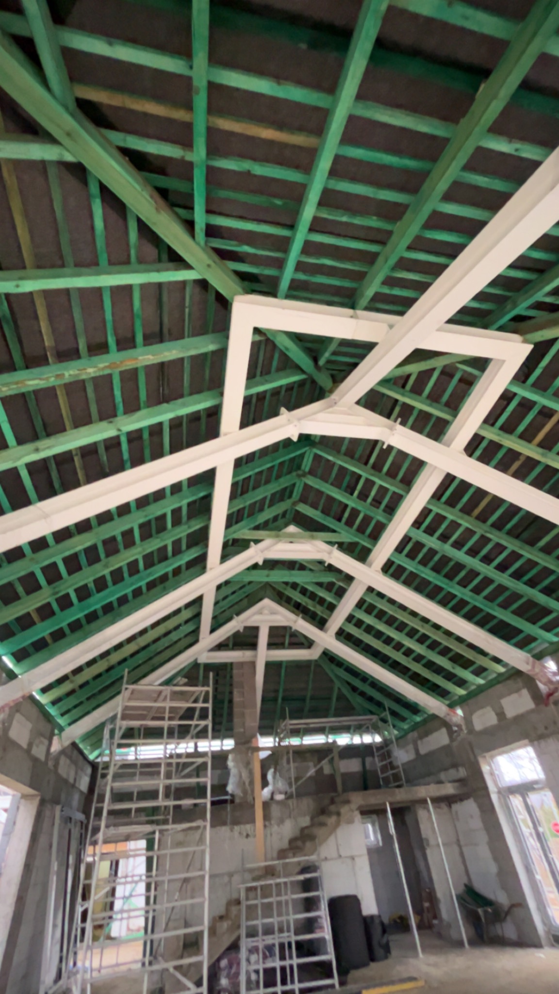 Wnętrze domu w trakcie budowy, widok na zieloną konstrukcję dachu z drewnianymi belkami i białymi elementami wzmacniającymi, rusztowania w tle.