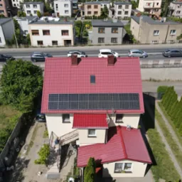 Widok z lotu ptaka na dom jednorodzinny z czerwoną dachówką i panelami fotowoltaicznymi na dachu, otoczony zielenią i zabudową sąsiedzką.