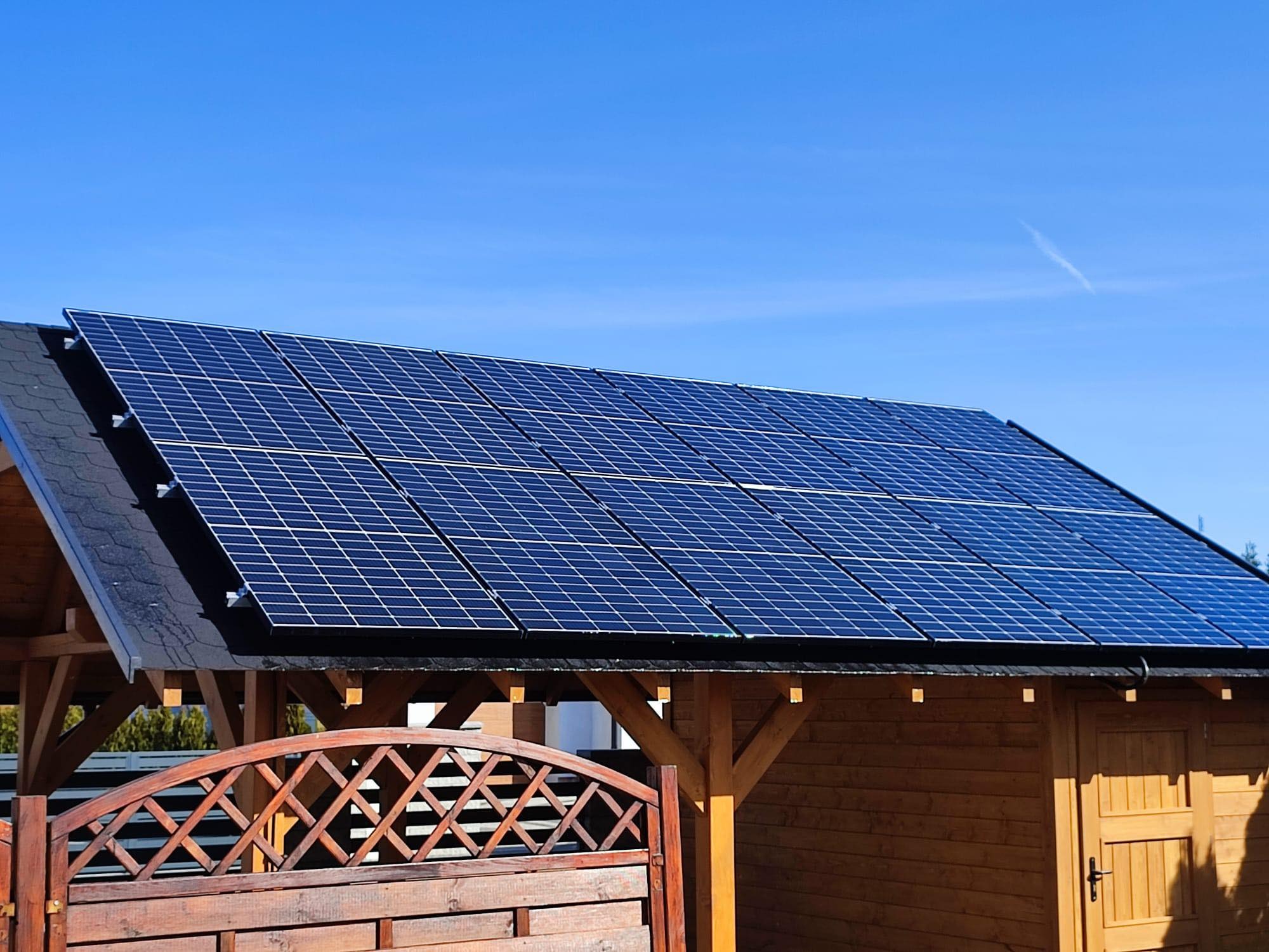 Panele słoneczne na dachu drewnianej altany w słoneczny dzień, widoczne drewniane elementy konstrukcyjne i ażurowa barierka.