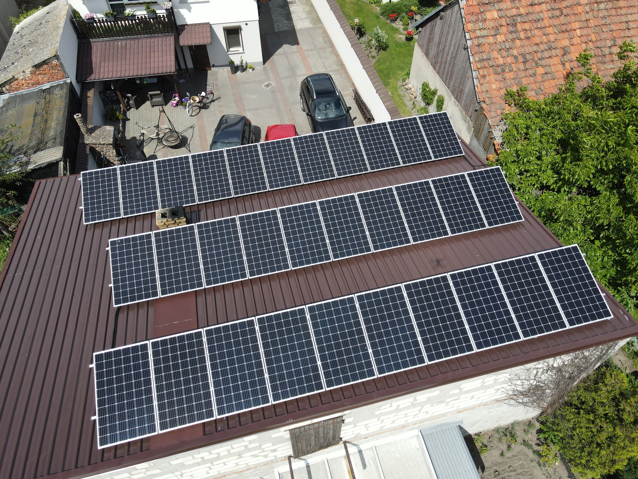 Widok z góry na brązowy dach z panelami słonecznymi; w tle podwórko z samochodami i zabudowaniami.