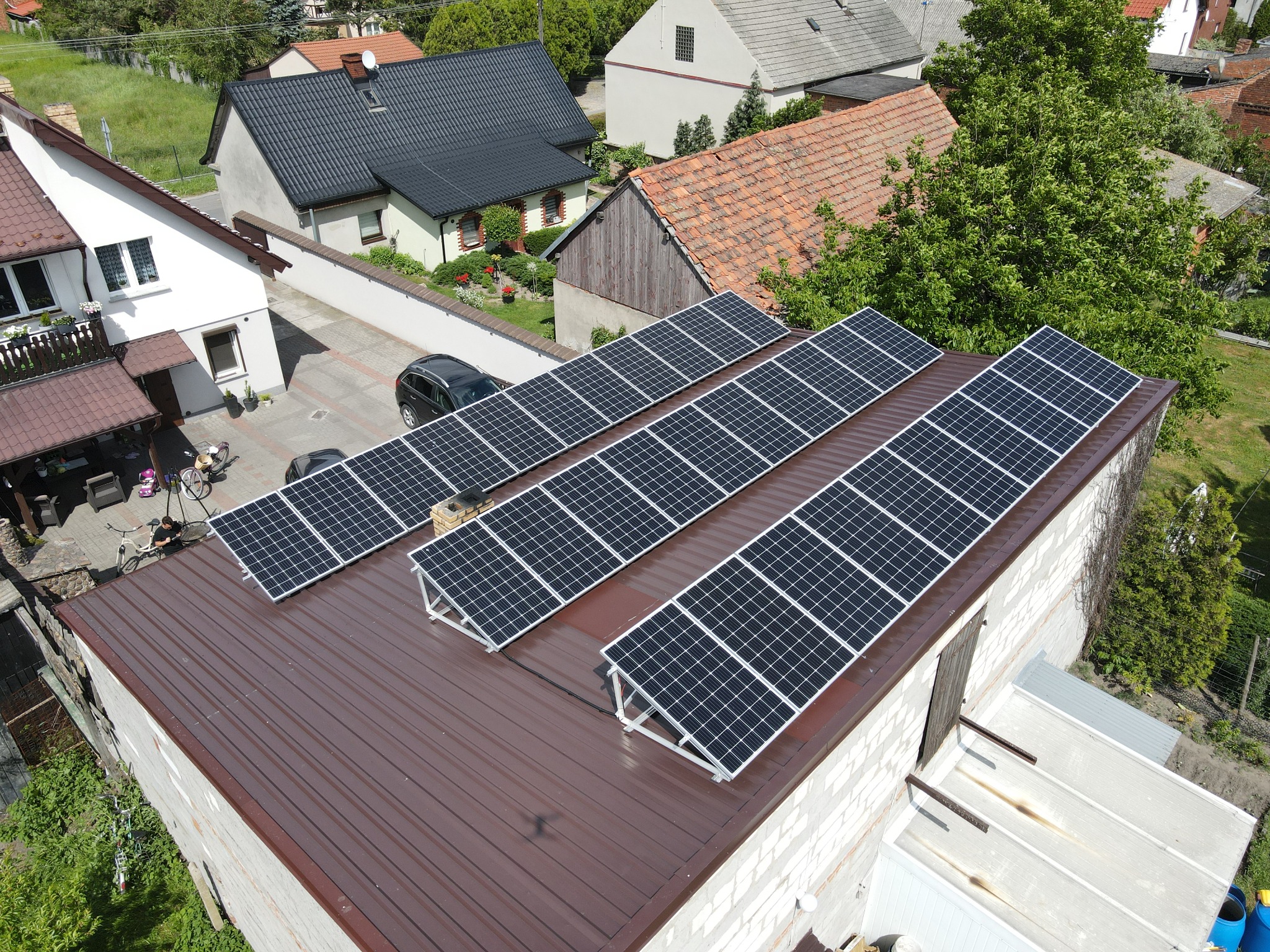 Widok z góry na panele słoneczne zamontowane na dachu budynku gospodarczego z brązowym dachem, w tle domy jednorodzinne i samochód na podjeździe.