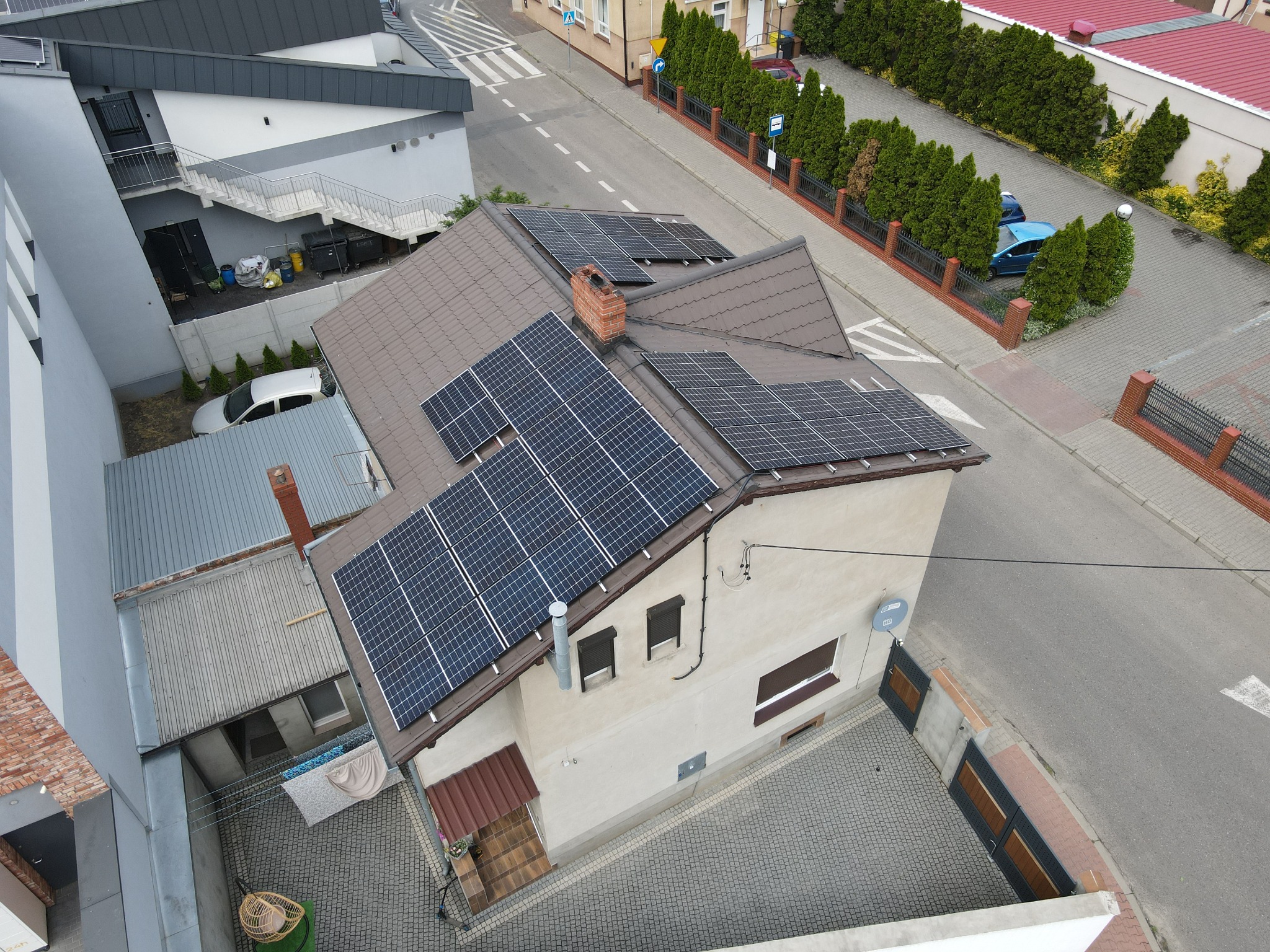 Widok z góry na dom jednorodzinny z brązowym dachem, na którym zamontowane są panele słoneczne; widoczny komin z czerwonej cegły, ulica i sąsiednie budynki.