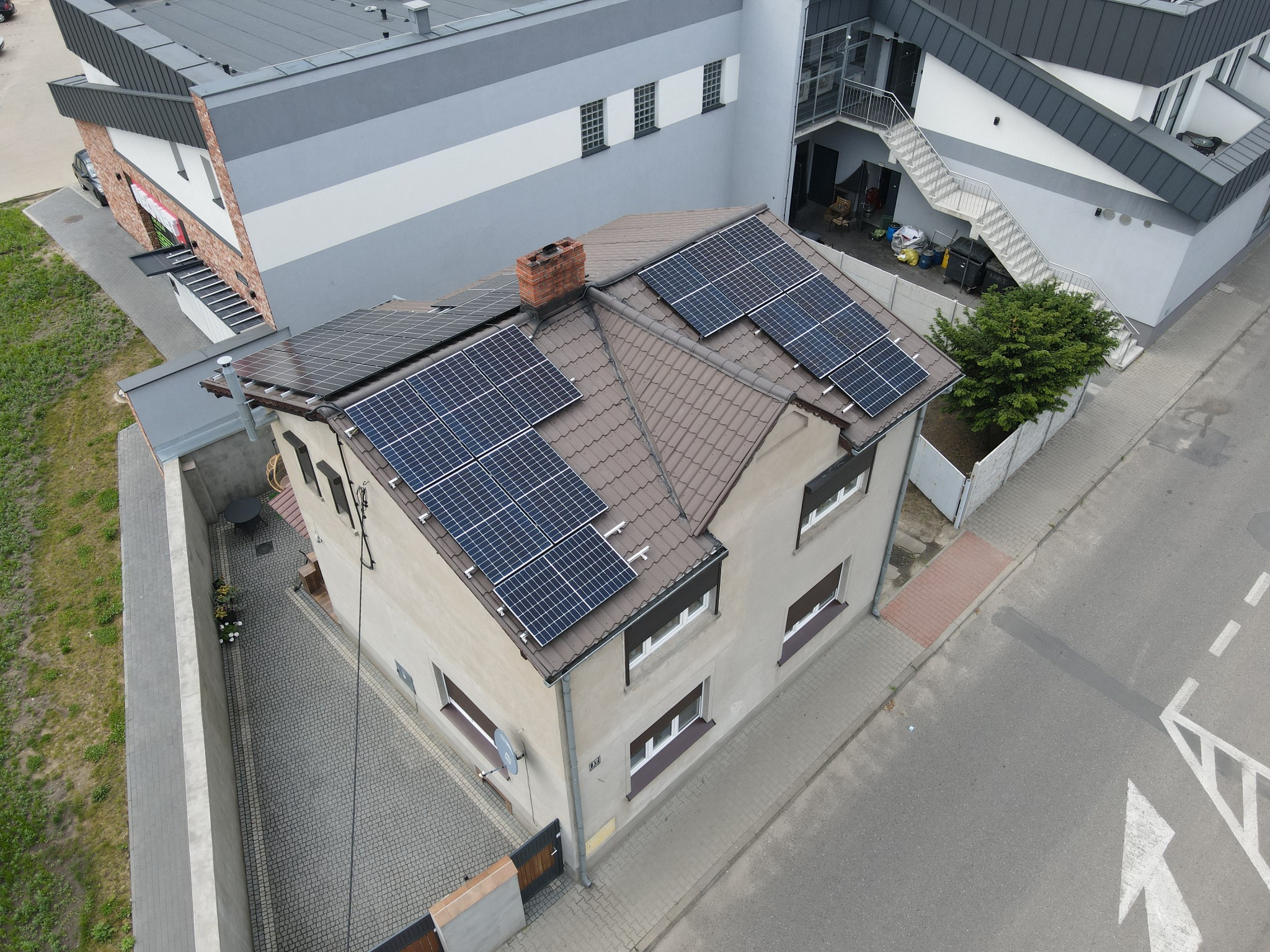 Widok z góry na dom jednorodzinny z brązową dachówką i panelami fotowoltaicznymi, z ceglanym kominem, obok nowoczesny budynek z zewnętrznymi schodami ewakuacyjnymi, małe drzewo i kostka brukowa.