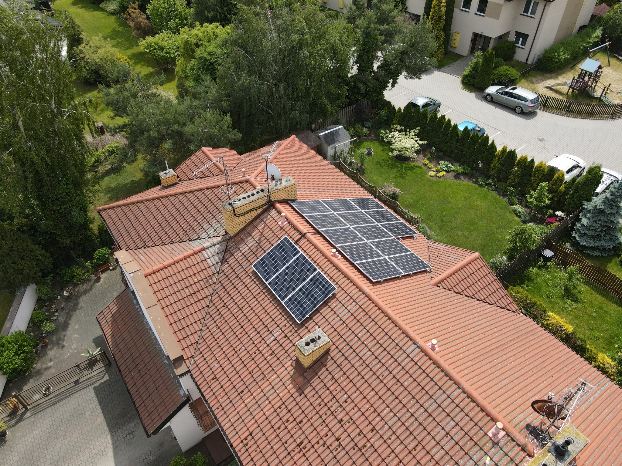 Widok z góry na dom z czerwoną dachówką, na której zamontowano panele słoneczne; widoczny fragment ogrodu i zabudowań w tle.
