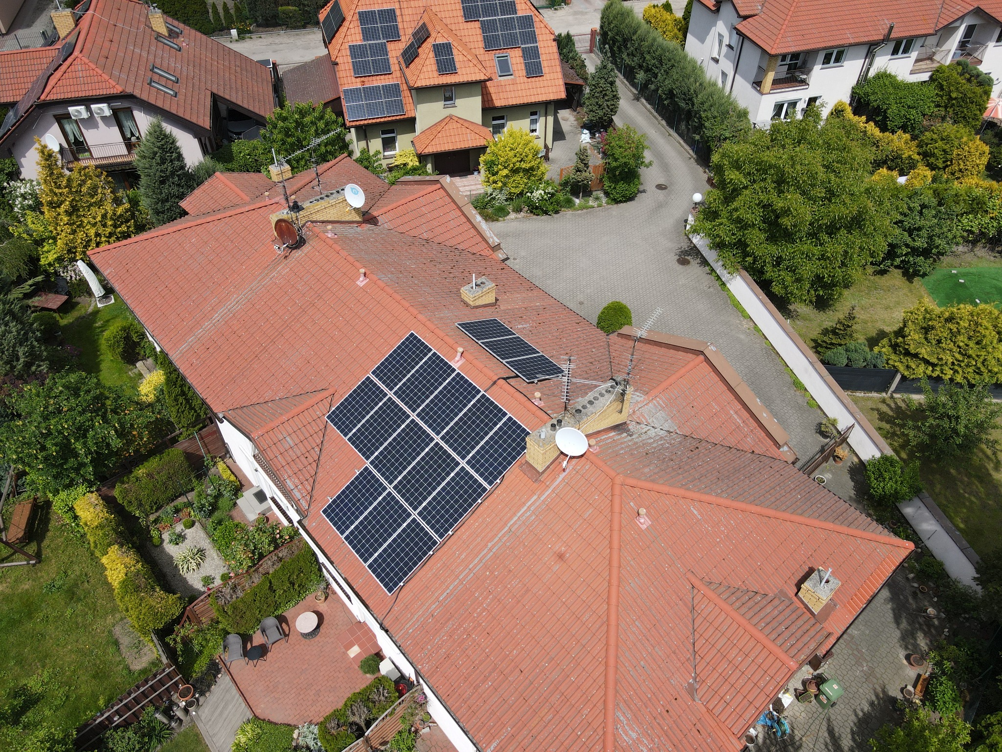 Widok z góry na dom z czerwoną dachówką, na której zamontowane są panele słoneczne, widoczne również anteny satelitarne i komin.