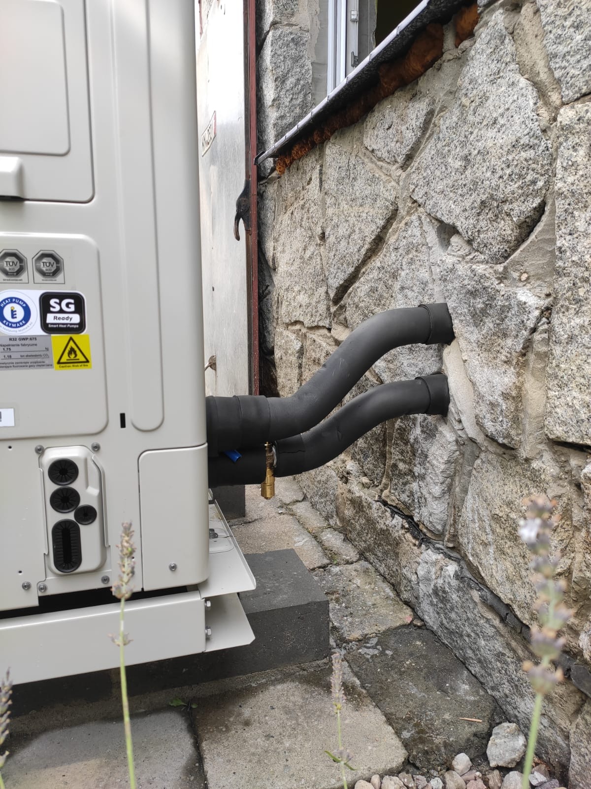 Zewnętrzna jednostka pompy ciepła zamontowana przy ścianie z kamienia, widoczne rury w izolacji termicznej wchodzące w otwór w ścianie oraz etykiety informacyjne na obudowie urządzenia.