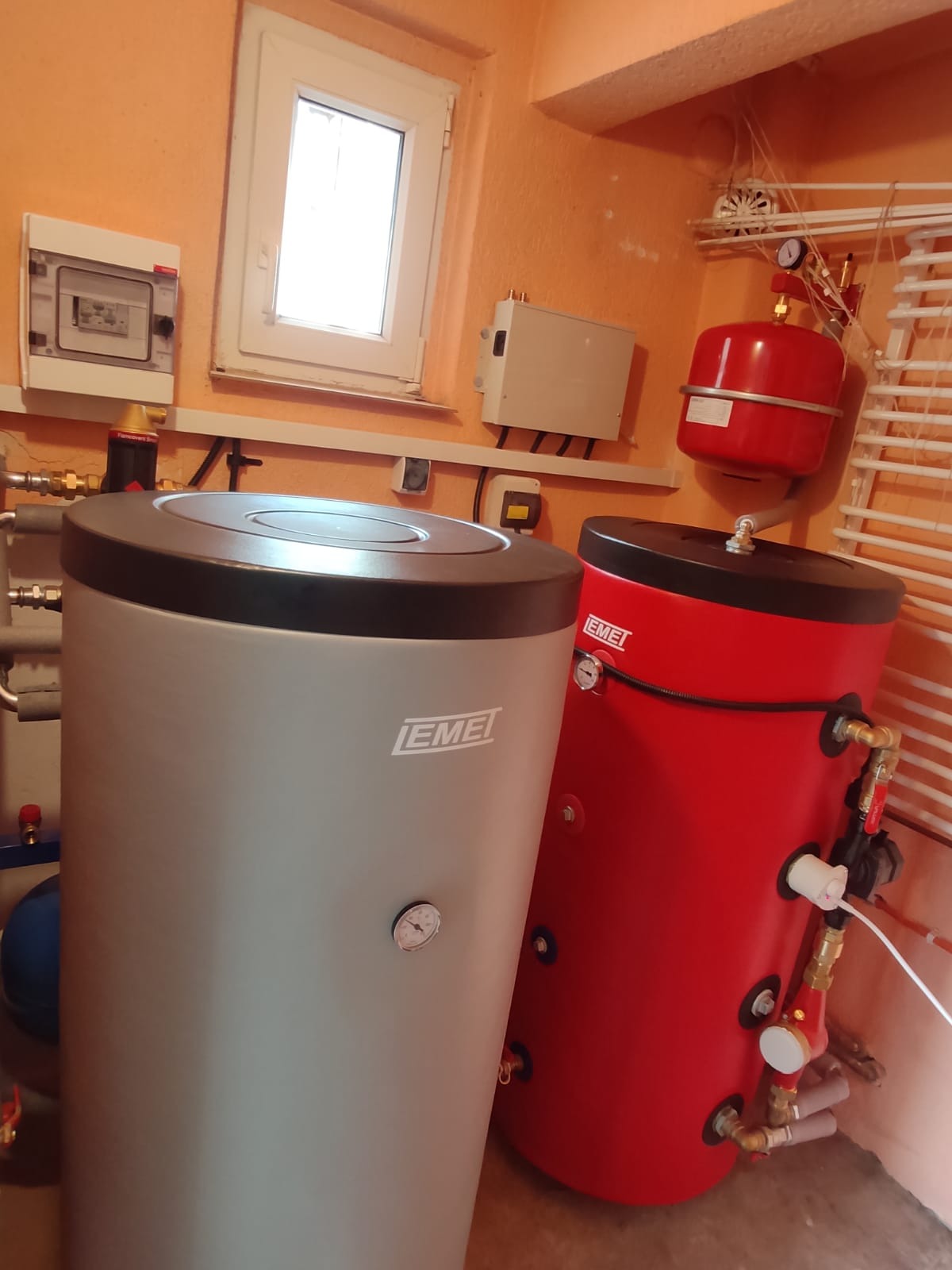 Szary i czerwony zbiornik akumulacyjny ciepła marki EMEI w kotłowni, widoczne rury, zawory i manometry, na ścianie okno i elementy instalacji elektrycznej.