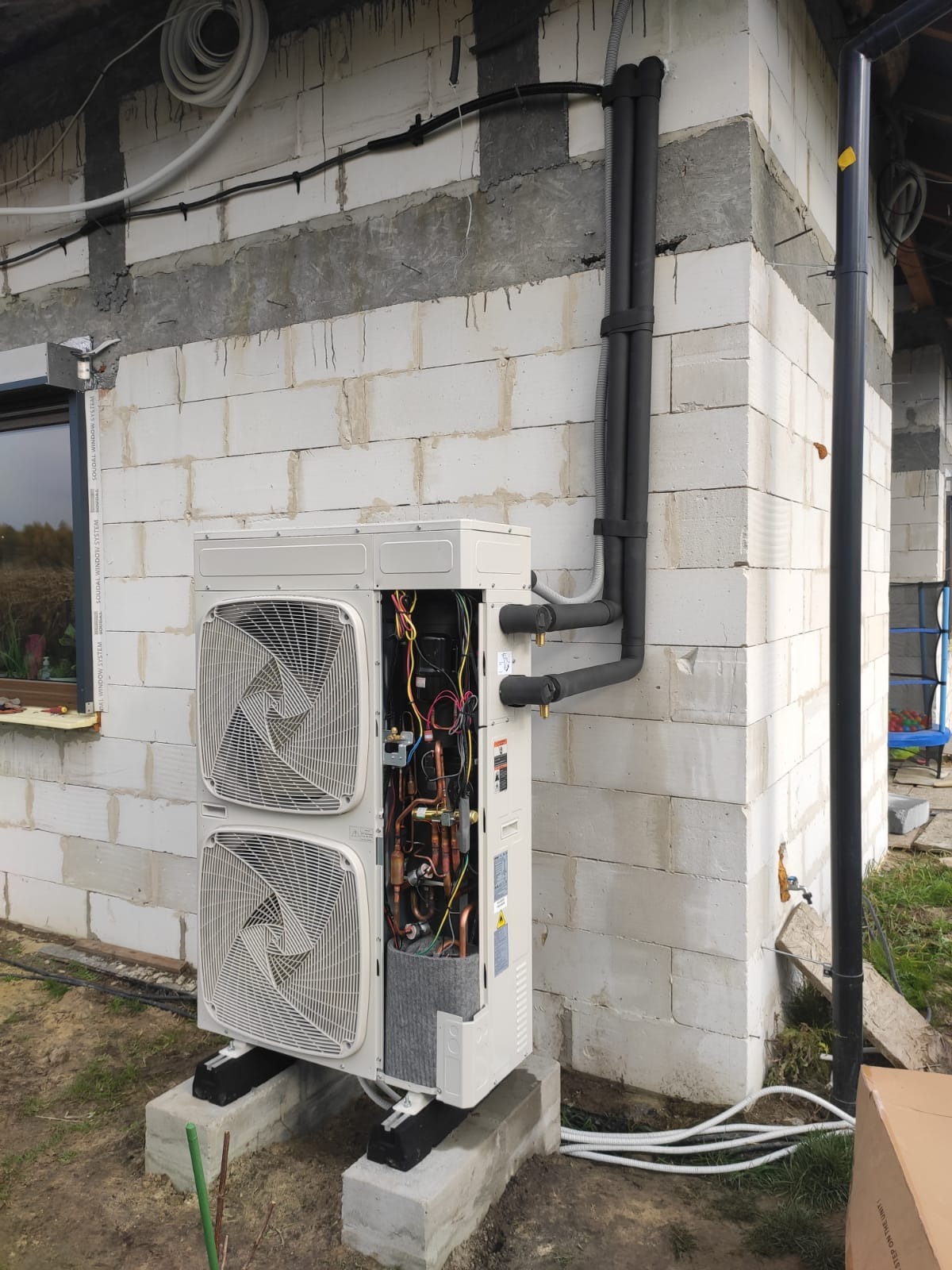 Zewnętrzna jednostka pompy ciepła typu split, z otwartą obudową ukazującą wewnętrzne komponenty, zamontowana na betonowych podstawach przy ścianie budynku z betonu komórkowego. Widoczne izolowane...