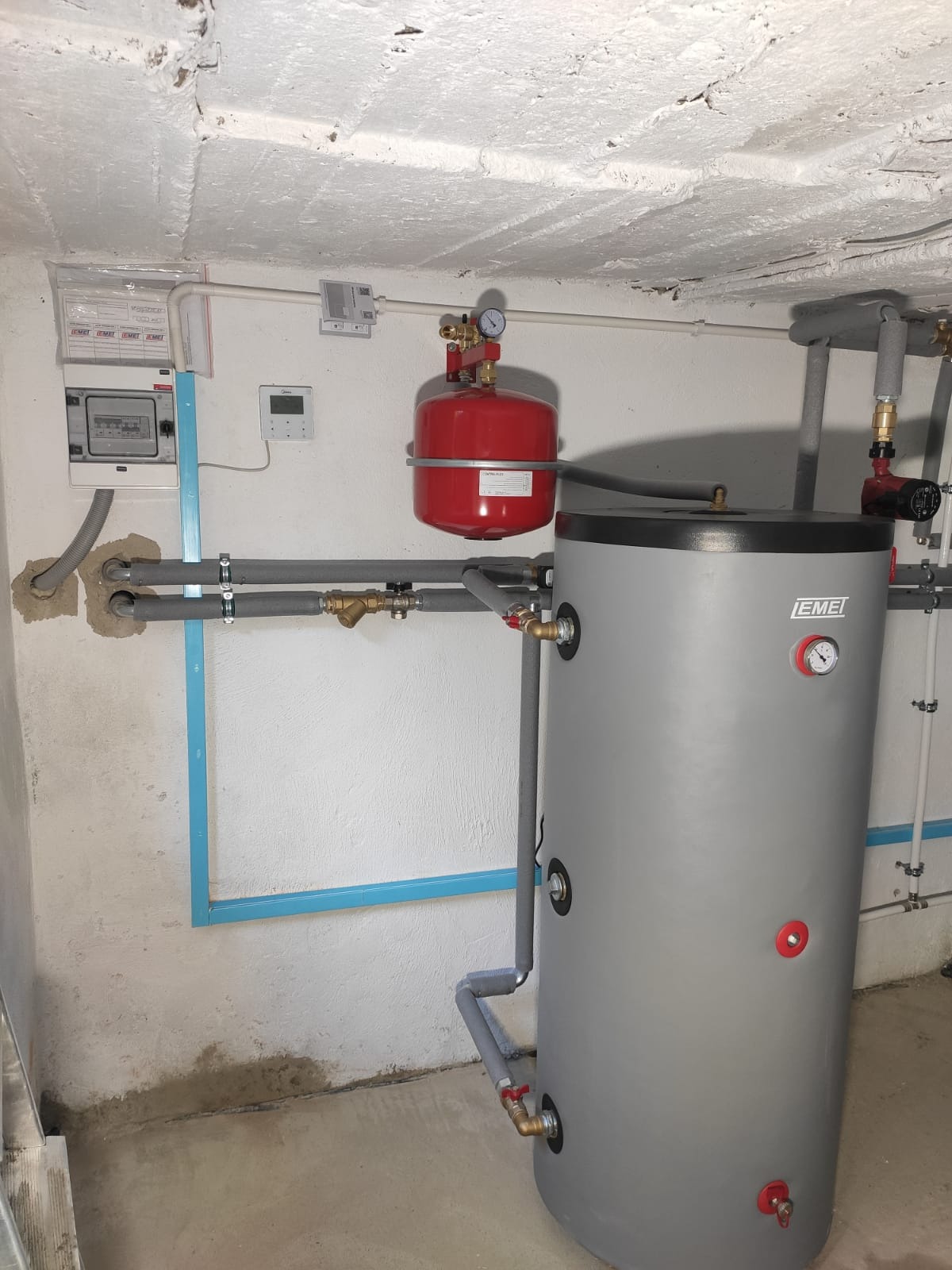 Szary zbiornik akumulacyjny ciepła EMEI z czerwonym naczyniem wzbiorczym, zamontowany w piwnicy z widocznymi rurami, zaworami i manometrami, na tle surowej ściany i sufitu.