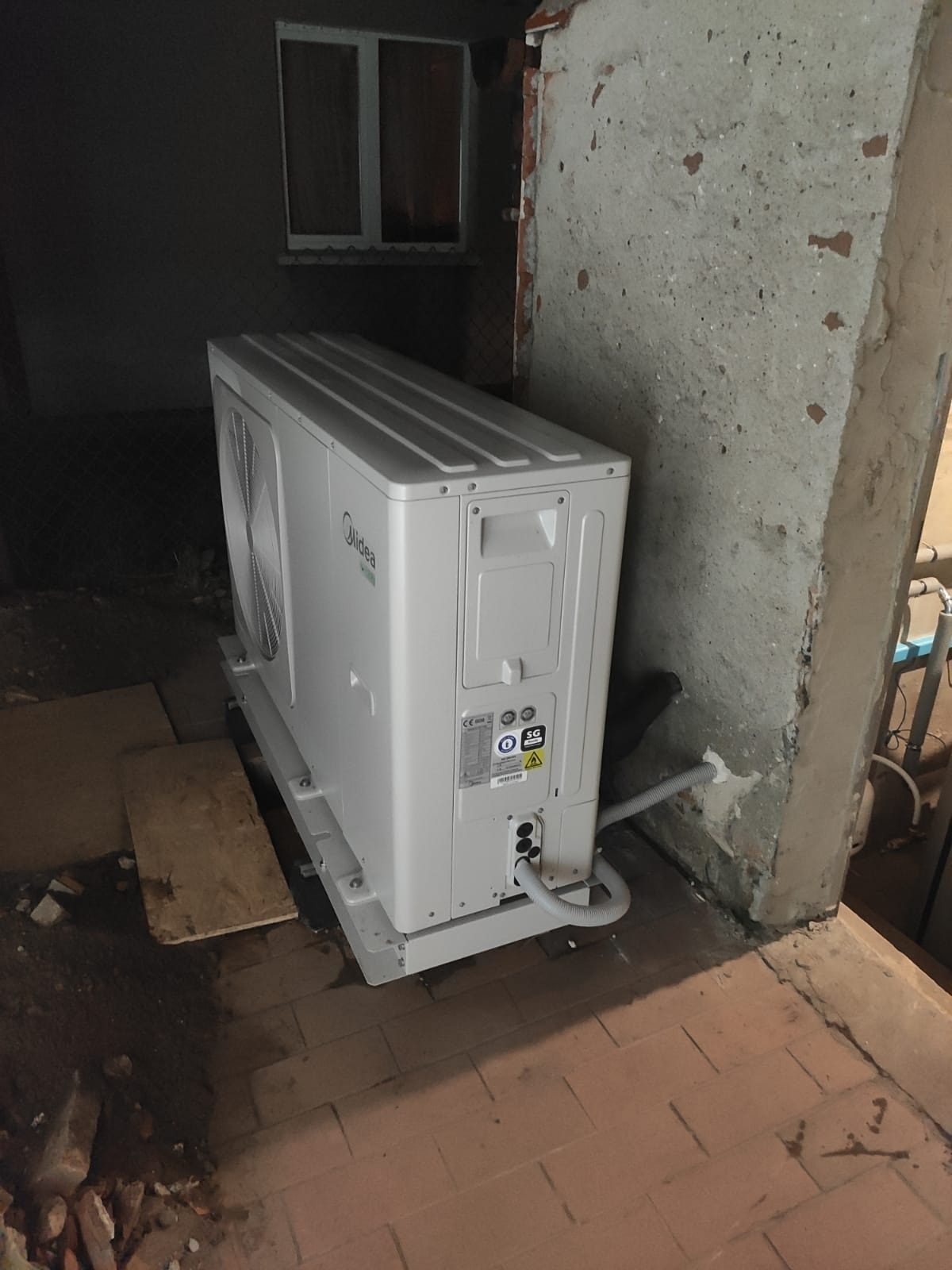 Zewnętrzna jednostka klimatyzacji Midea na metalowej podstawie, z widocznymi przewodami, zamontowana przy starej, betonowej ścianie w piwnicy, z małym oknem w tle.