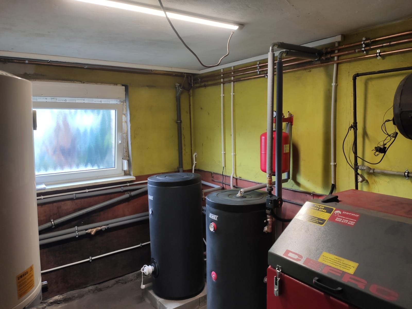 Kotłownia z dwoma czarnymi zbiornikami akumulacyjnymi EMEI, czerwonym naczyniem wzbiorczym i piecem Defro z naklejkami informacyjnymi, rurami miedzianymi i oświetleniem jarzeniowym.