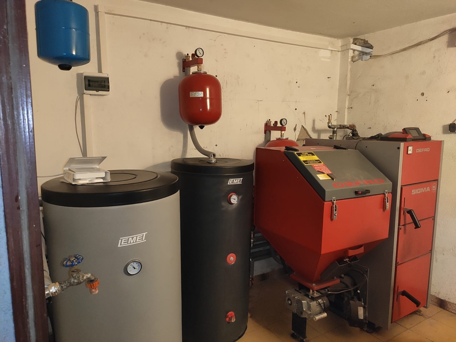 Kotłownia z kotłem na paliwo stałe Defro Sigma 5, dwoma zbiornikami buforowymi IEMEi z osprzętem oraz naczyniami wzbiorczymi w kolorach czerwonym i niebieskim, zamontowanymi na ścianie