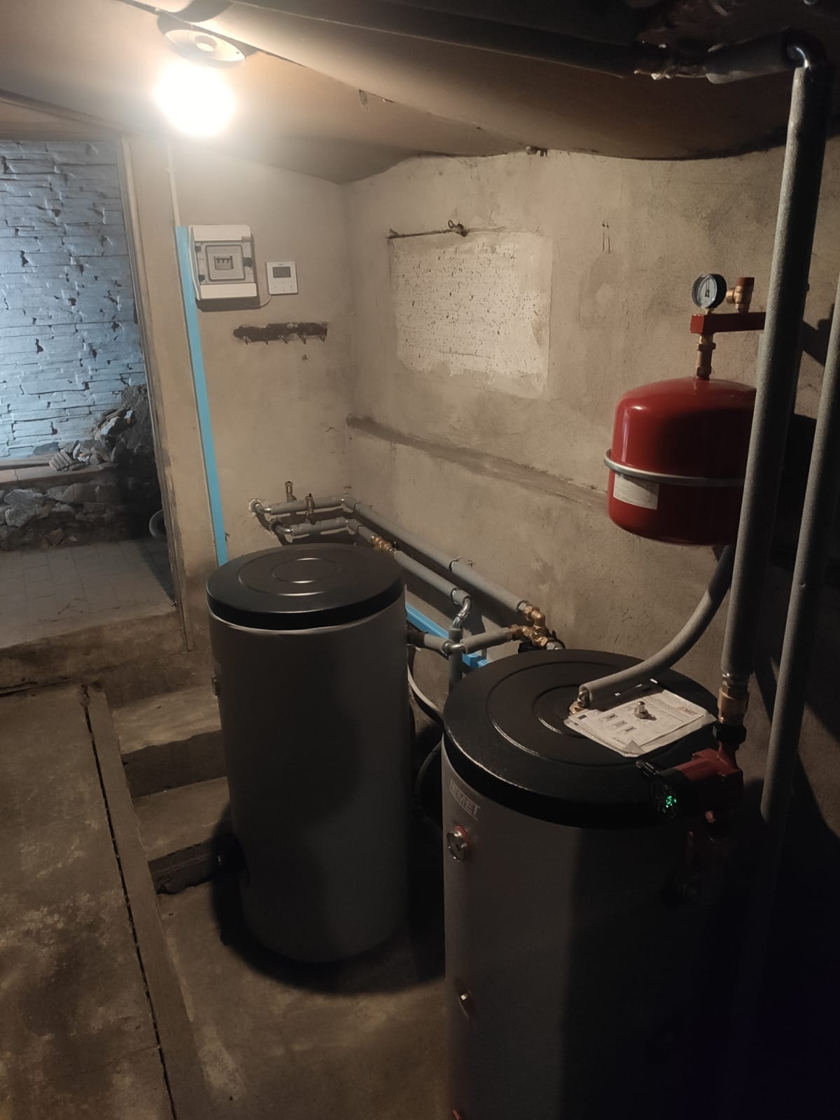 Dwa szare zbiorniki akumulacyjne w piwnicy, z czerwonym zbiornikiem wyrównawczym i instalacją rurową, oświetlone lampą sufitową. Widoczny fragment ściany z instalacją elektryczną.