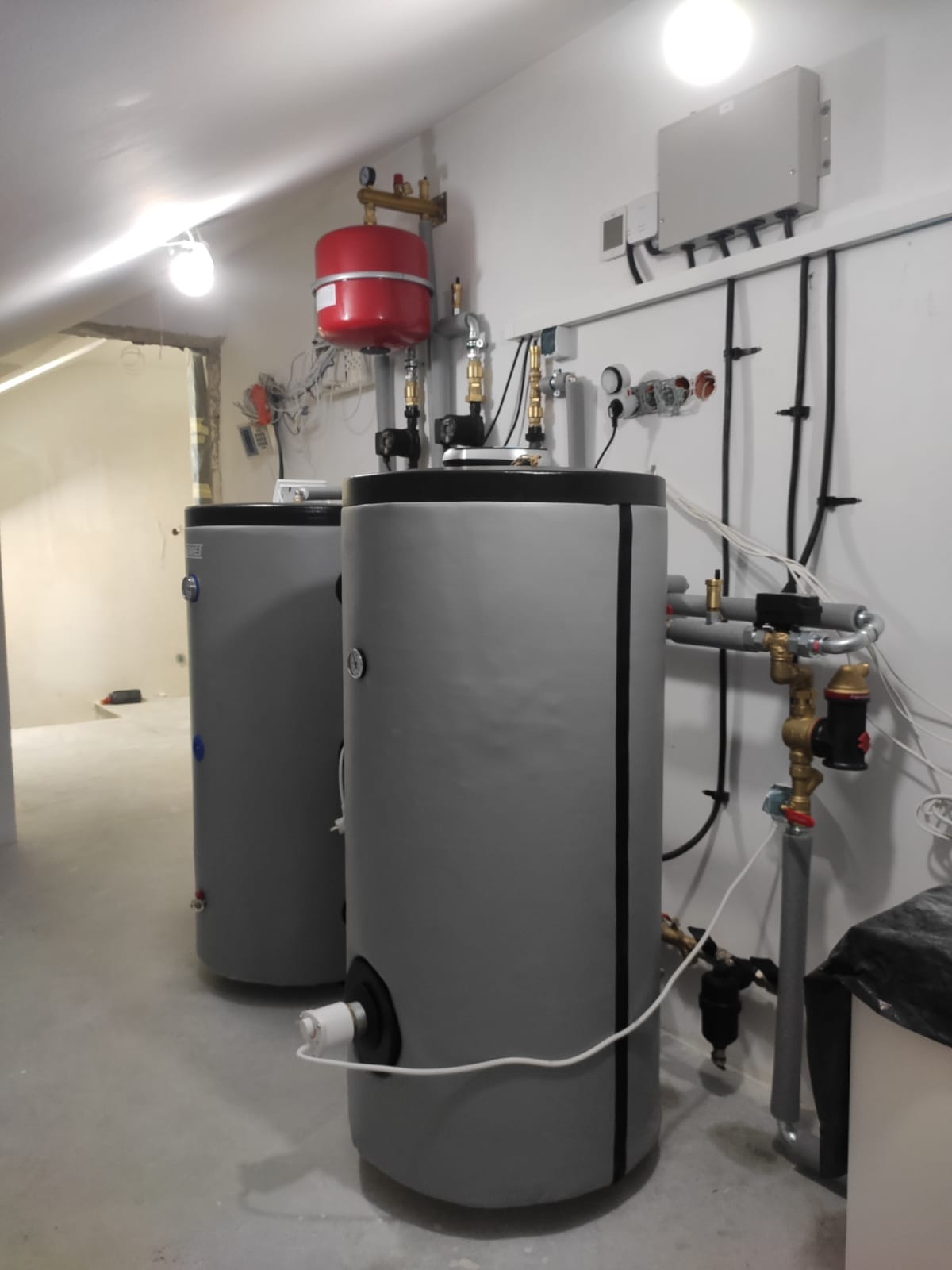 Dwa szare zbiorniki akumulacyjne ciepła z czerwoną instalacją nad nimi, zamontowane w pomieszczeniu z widocznymi przewodami i elementami instalacji na ścianie.
