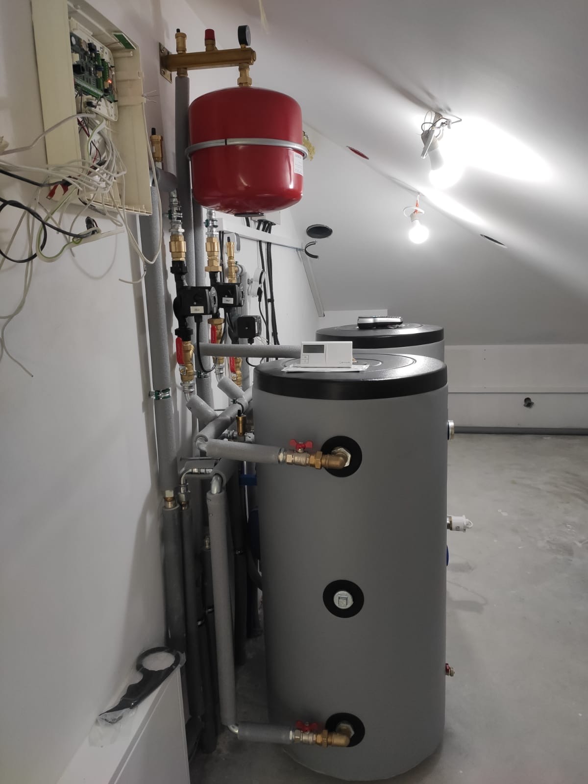 Instalacja dwóch szarych zbiorników akumulacyjnych z rurami i czerwonym naczyniem przeponowym w kotłowni w Zielonej Górze, widoczna instalacja elektryczna na ścianie.