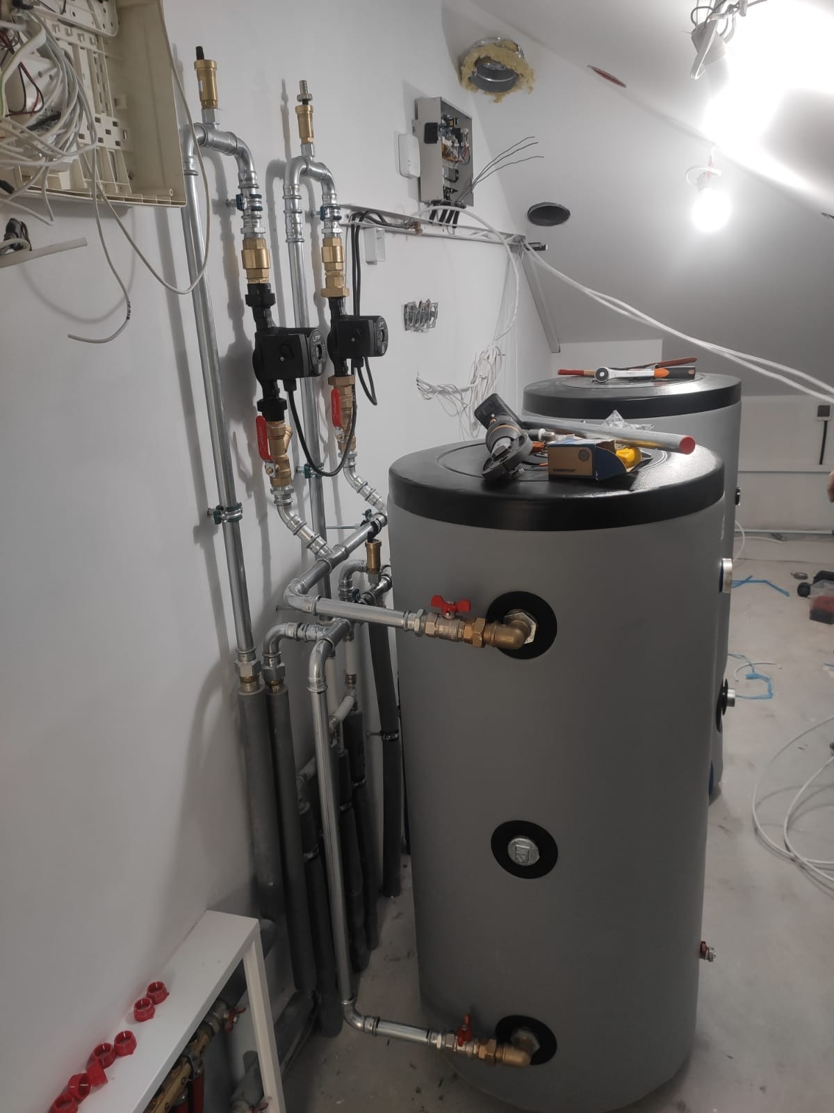 Dwa szare zbiorniki akumulacyjne z podłączonymi rurami i zaworami w pomieszczeniu na poddaszu, widoczne narzędzia na górze zbiornika, instalacja w trakcie montażu.