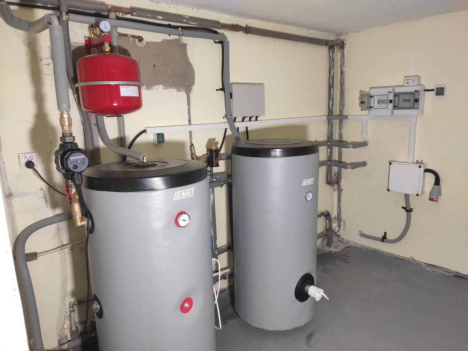Dwa szare zbiorniki buforowe EMEI w kotłowni, z czerwoną membraną i pompą obiegową Onnline, instalacja rurowa pod sufitem, szara podłoga