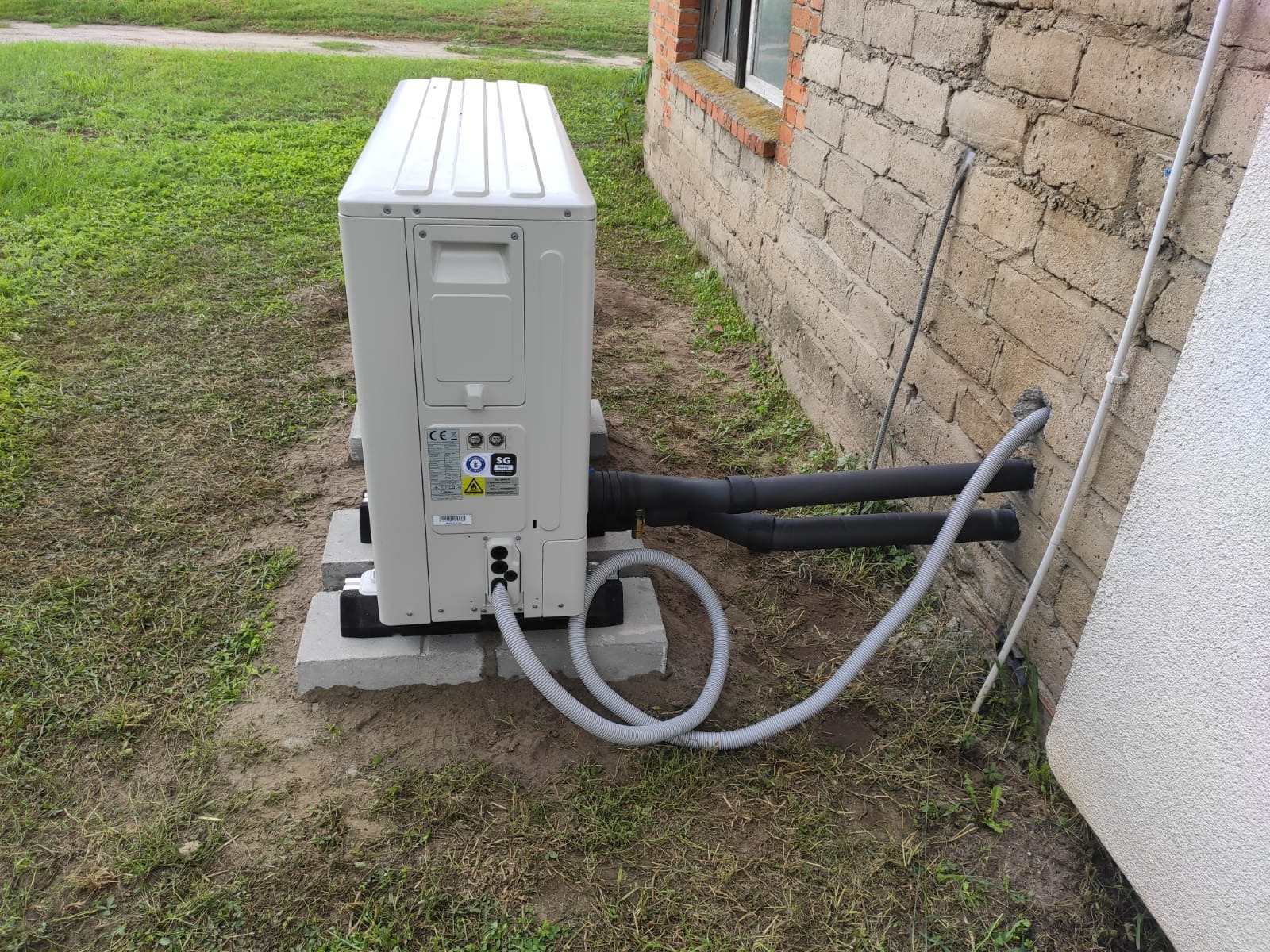 Zewnętrzna jednostka pompy ciepła, zamontowana na betonowych podstawach przy ścianie budynku z widocznymi rurami przyłączeniowymi i przewodami w peszlu.