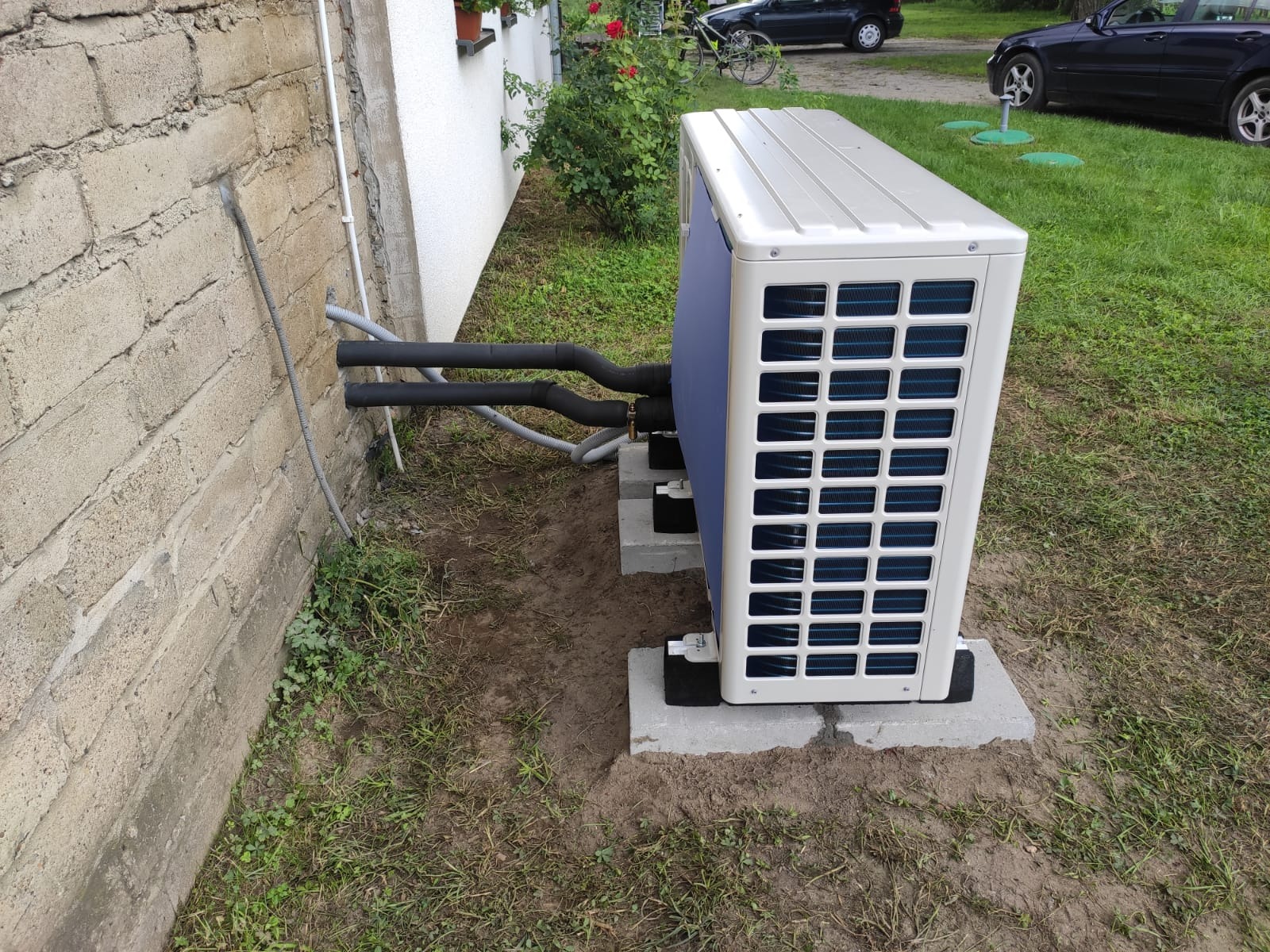Zewnętrzna jednostka pompy ciepła zamontowana na betonowych podstawach przy ścianie budynku z widocznymi izolowanymi rurami przyłączeniowymi.