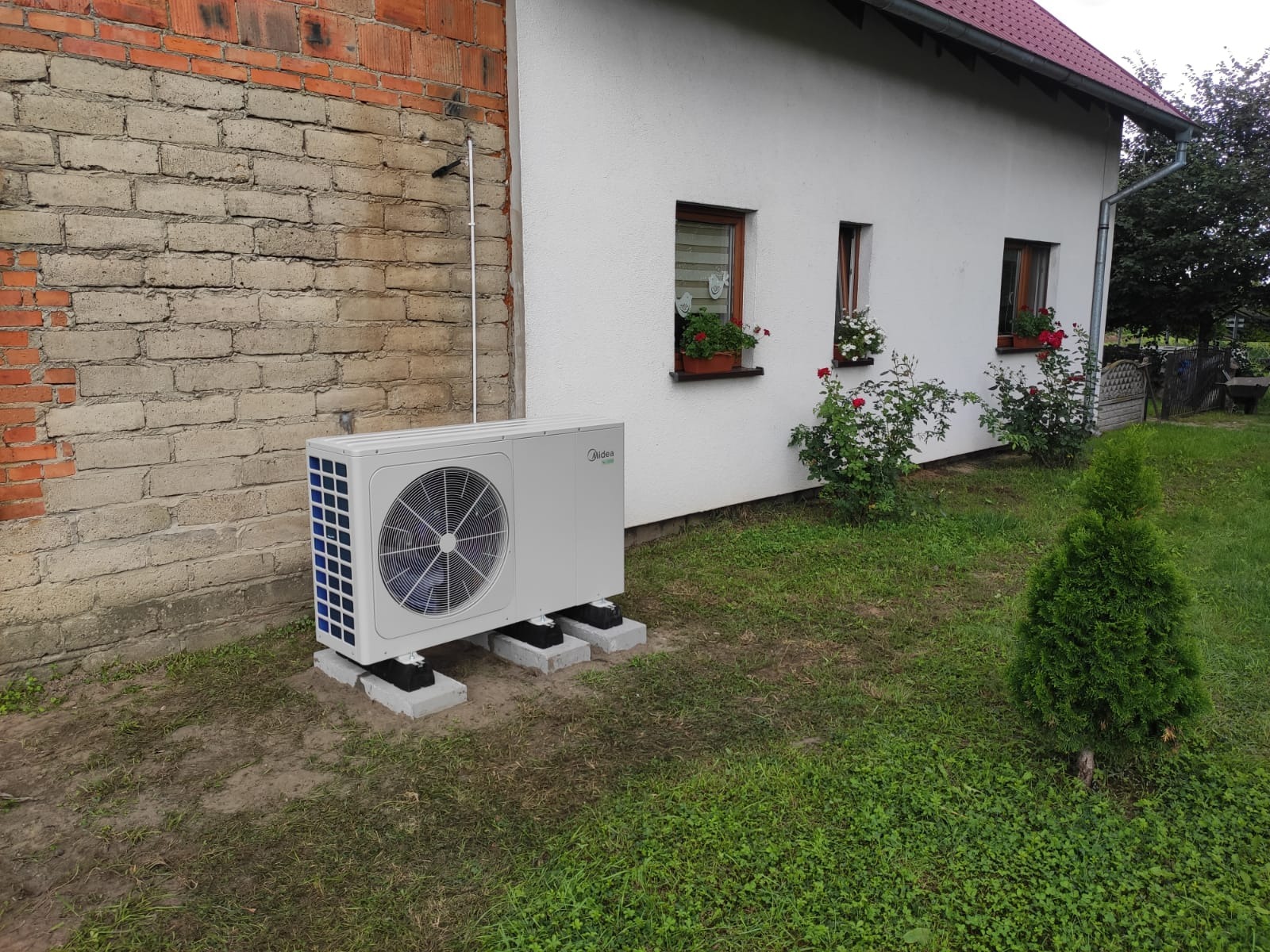 Zewnętrzna jednostka pompy ciepła Midea zamontowana na betonowych bloczkach przy ścianie domu, z widocznymi kwiatami w oknach i krzewami róż w ogrodzie.