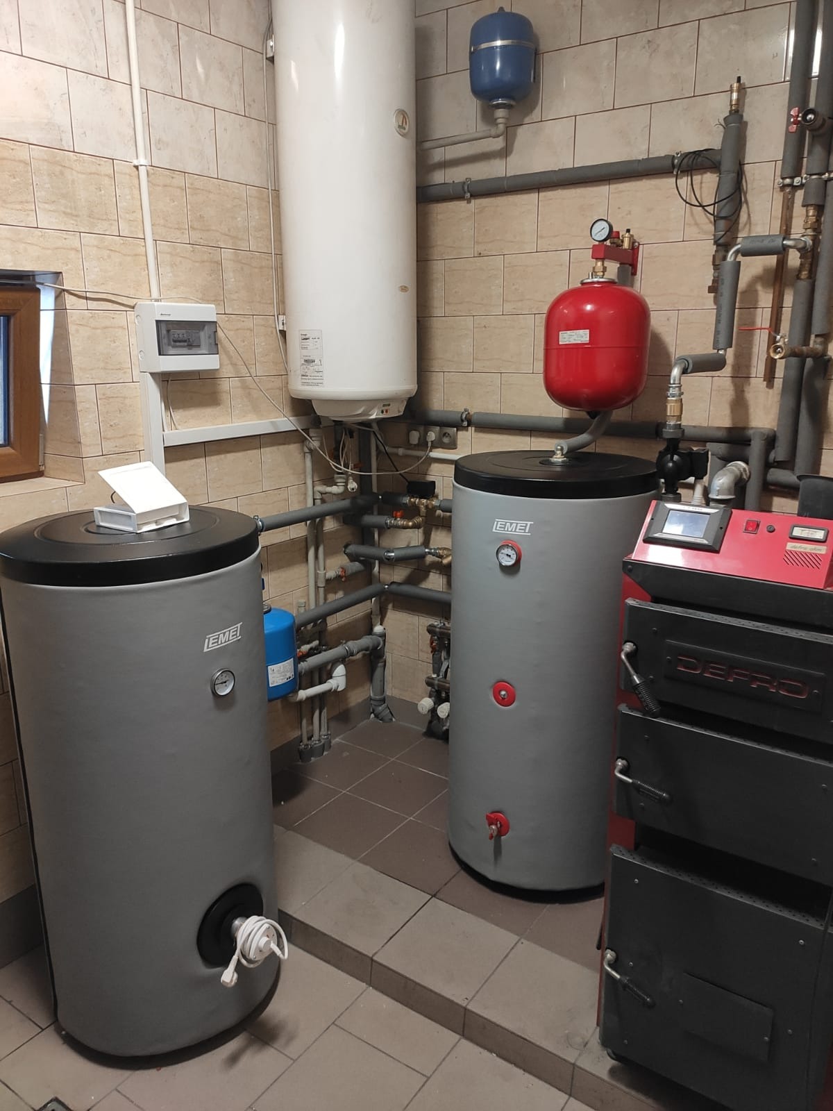Kotłownia z dwoma szarymi zbiornikami EMEI, białym bojlerem, czerwonym zbiornikiem wyrównawczym i czarnym piecem Defro, widoczne liczne rury i instalacje, całość na tle ceramicznej okładziny ściennej.