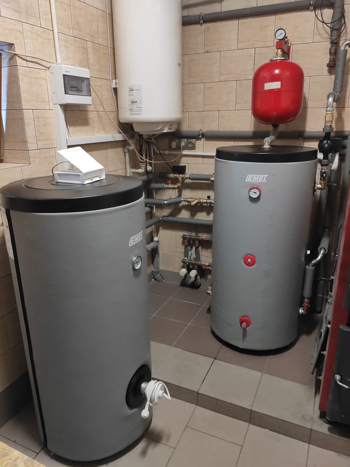 Dwa szare zasobniki ciepłej wody użytkowej marki Emei, z widocznymi manometrami i zaworami, zainstalowane w kotłowni z beżowymi płytkami ceramicznymi na ścianach i podłodze; dodatkowo widoczny...