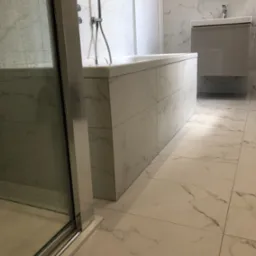 Wykończona łazienka z wanną obudowaną płytkami imitującymi marmur, szklaną kabiną prysznicową i szafką z umywalką w odcieniach szarości, całość utrzymana w jasnej kolorystyce.