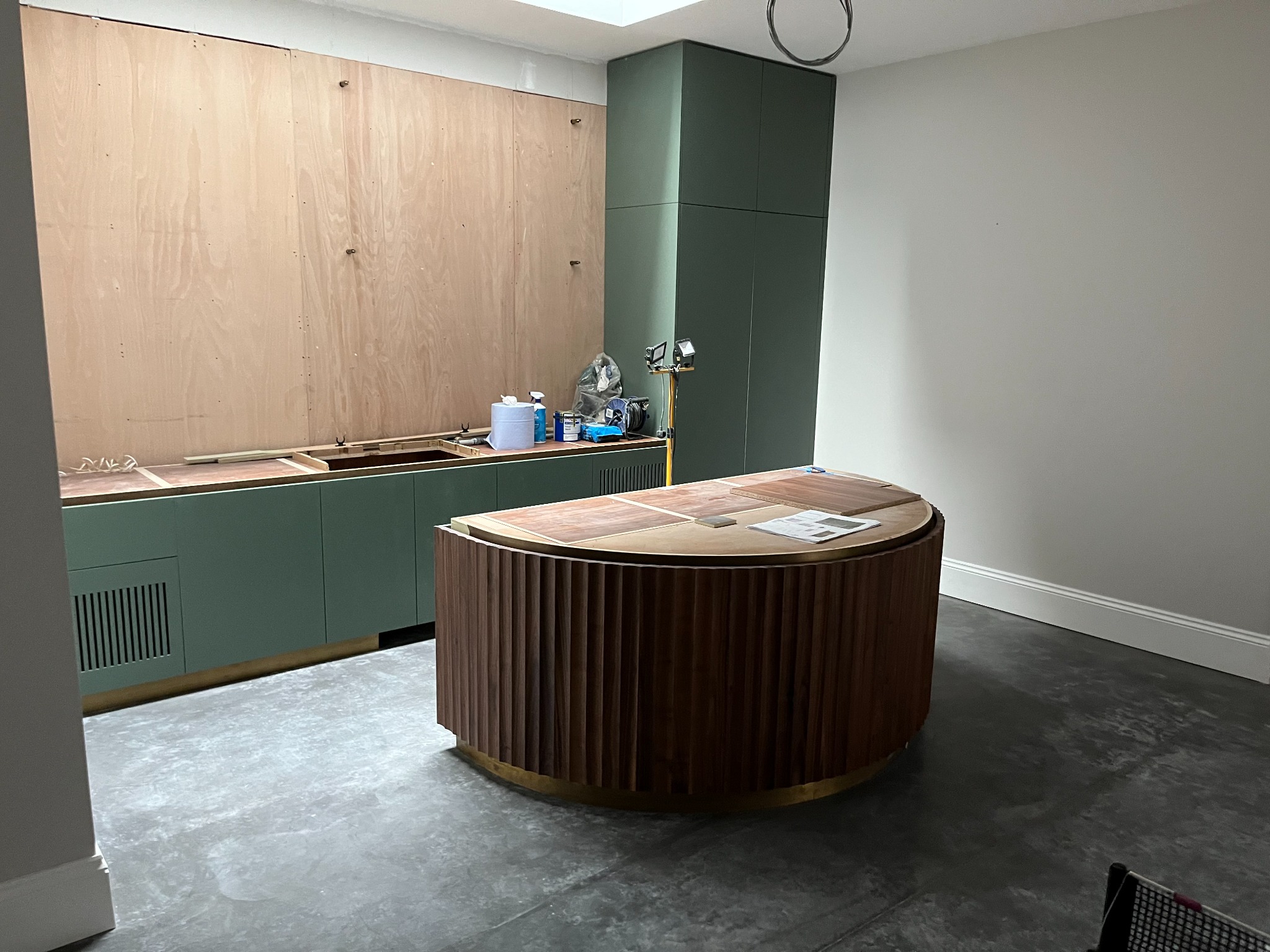 Wnętrze kuchni w trakcie montażu: zielone szafki dolne z drewnianym blatem, drewniana wyspa z ryflowanym wykończeniem, oraz wysoka zabudowa w kolorze zieleni, podświetlona lampą budowlaną.