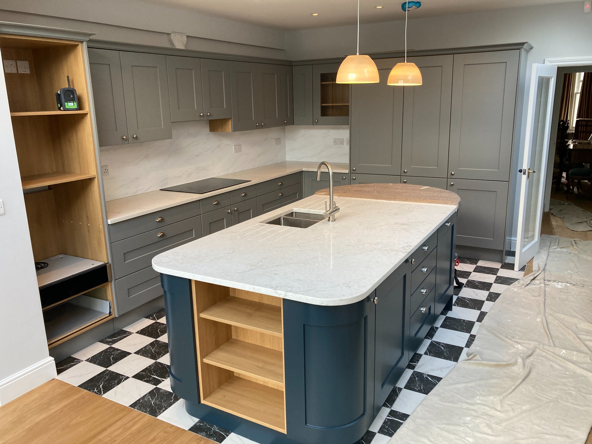 Wnętrze kuchni z szarymi szafkami i wyspą z niebieskimi frontami i marmurowym blatem, czarno-biała podłoga w szachownicę, widoczne lampy wiszące i częściowo zakryta podłoga folią ochronną.