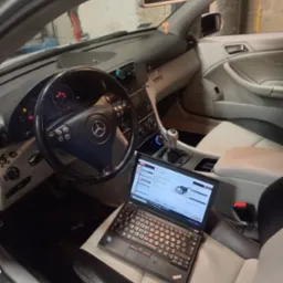 Wnętrze samochodu Mercedes z laptopem na siedzeniu pasażera, wyświetlającym interfejs diagnostyczny, deska rozdzielcza z włączonymi kontrolkami, widoczna kierownica z logo Mercedes.