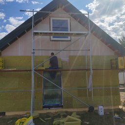 USŁUGI OGÓLNOBUDOWLANE PAWEŁ TELEGA - Ocieplanie elewacji domu jednorodzinnego z użyciem rusztowania. Widoczne płyty izolacyjne z wełny mineralnej, częściowo zamontowane. Prace w toku, słoneczny dzień.