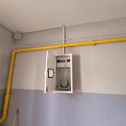 USŁUGI OGÓLNOBUDOWLANE PAWEŁ TELEGA - Otwarta skrzynka z instalacją elektryczną na dwukolorowej ścianie, z żółtą rurą gazową biegnącą wzdłuż sufitu. Widoczne przewody i elementy instalacji.