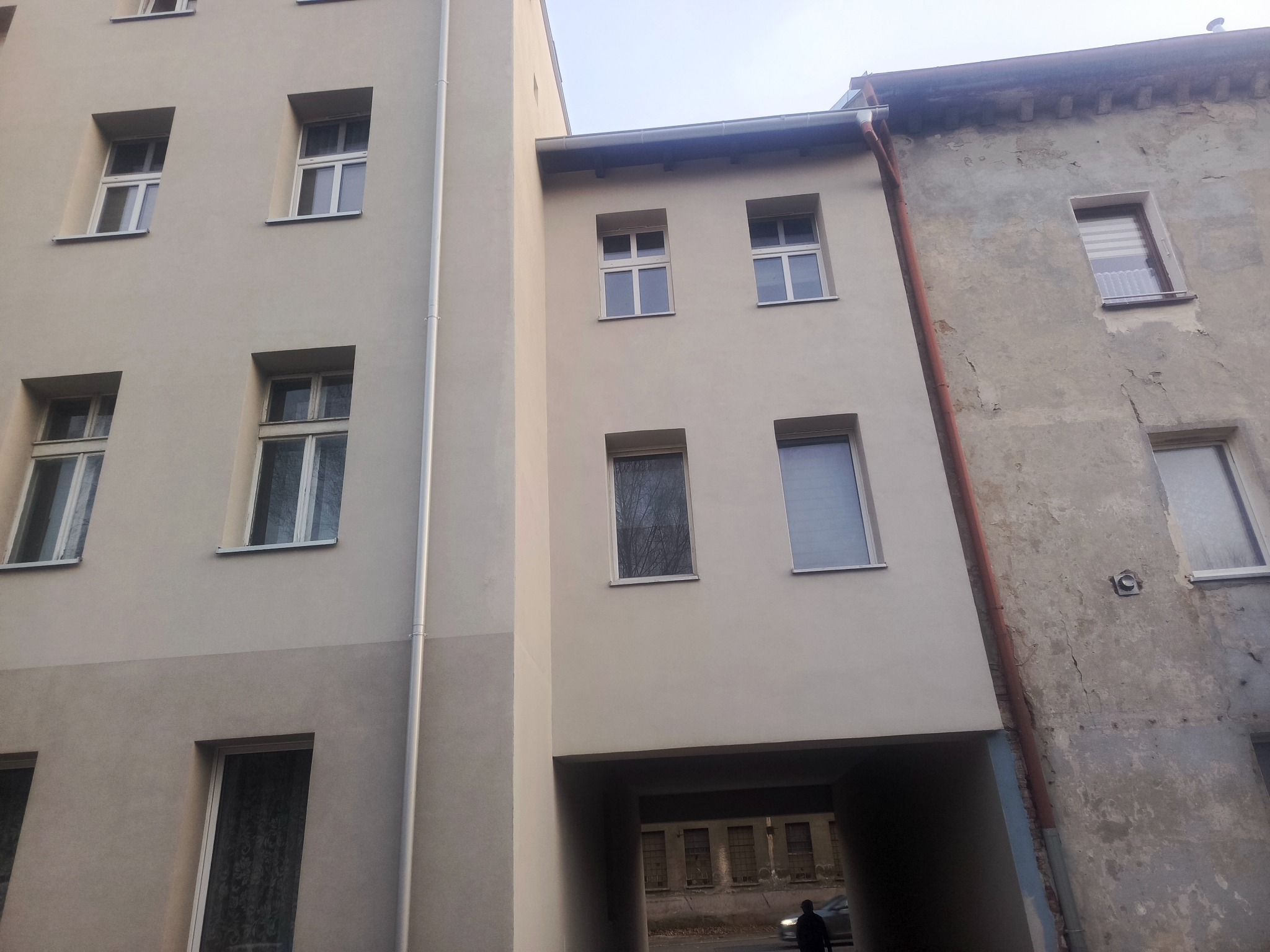 Odnowiona elewacja budynku z jasną fasadą i oknami, kontrastująca ze starą, szarą ścianą sąsiedniego budynku. Widoczny przejazd bramowy.