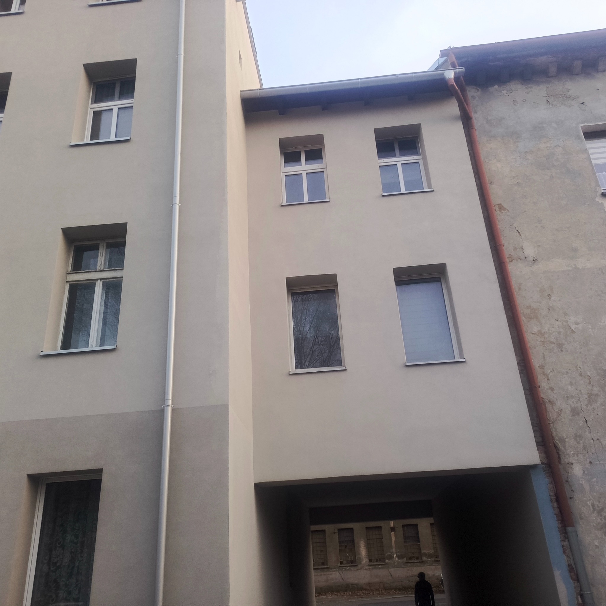 Odnowiona elewacja budynku z jasną farbą, widoczne okna i rynna. Fragment starej, nieodnowionej elewacji po prawej stronie. Przejście bramowe w budynku.