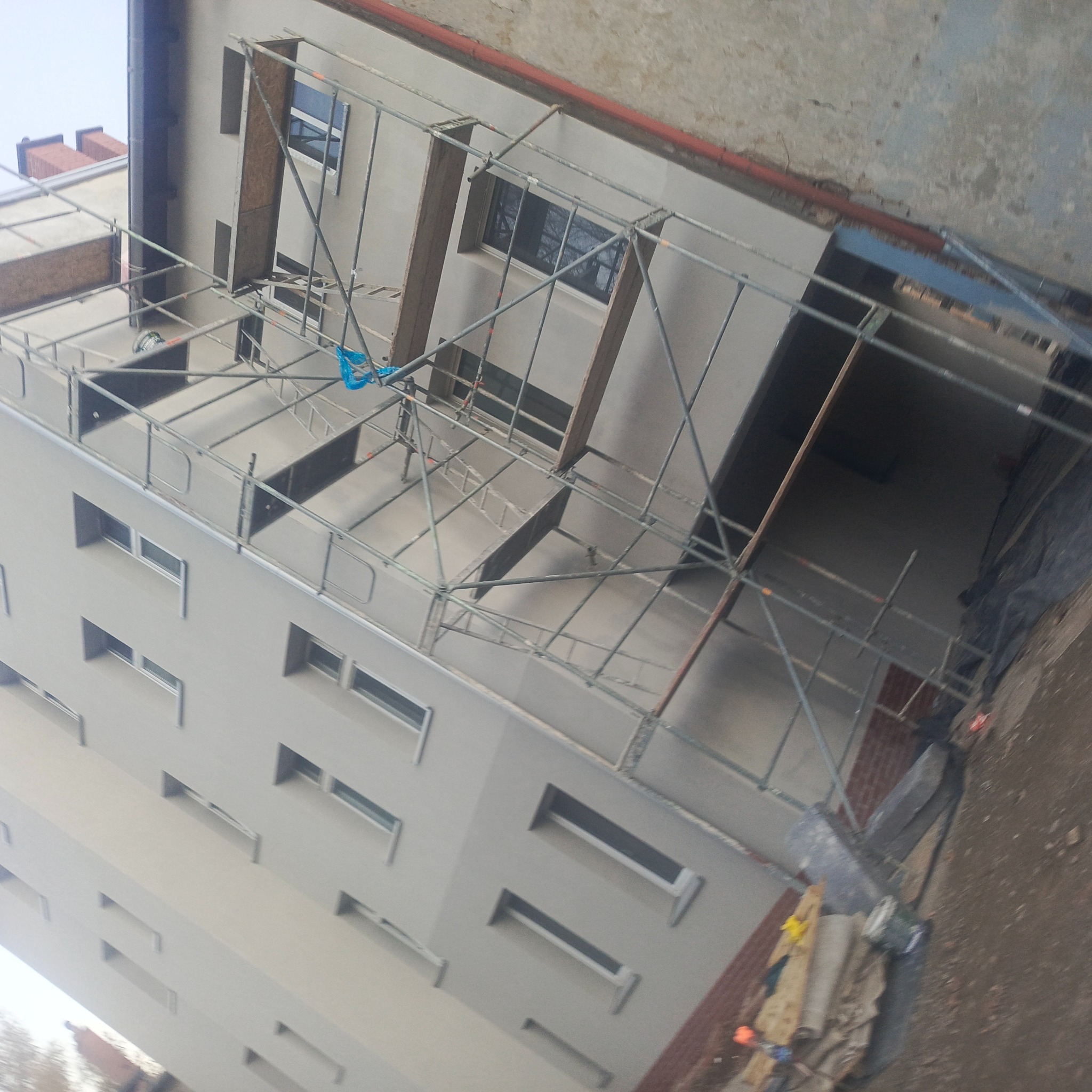 Elewacja budynku w trakcie malowania, widoczne rusztowania i świeżo pomalowana ściana. Ujęcie z dołu, podkreślające wysokość budynku i postęp prac.