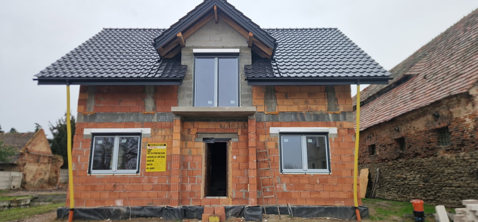 Surowy stan domu jednorodzinnego z czerwonej cegły z zamontowanymi oknami i czarnym dachem. Widoczna drabina i żółta rura. W tle budynek gospodarczy.