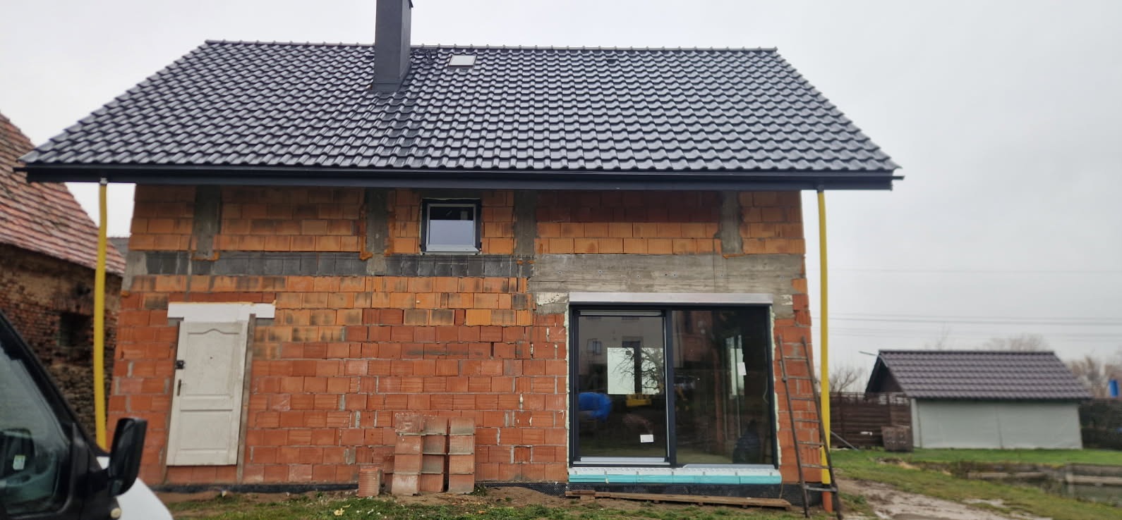 Surowy stan budynku w trakcie budowy: czerwona cegła, czarny dach z kominem, okno, drzwi. Widoczne elementy rusztowania i drabina. Tło: pochmurne niebo, budynek gospodarczy.