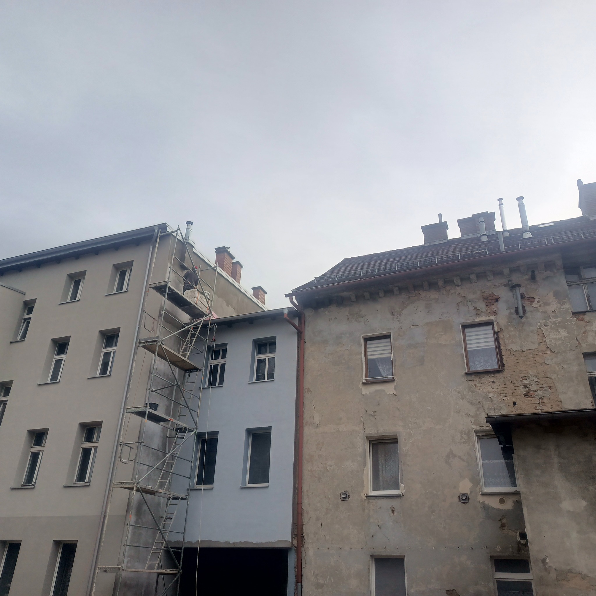 Budynek przed i po renowacji elewacji. Po lewej odnowiona fasada w jasnych odcieniach, po prawej stara, zniszczona elewacja. Widoczne rusztowania.