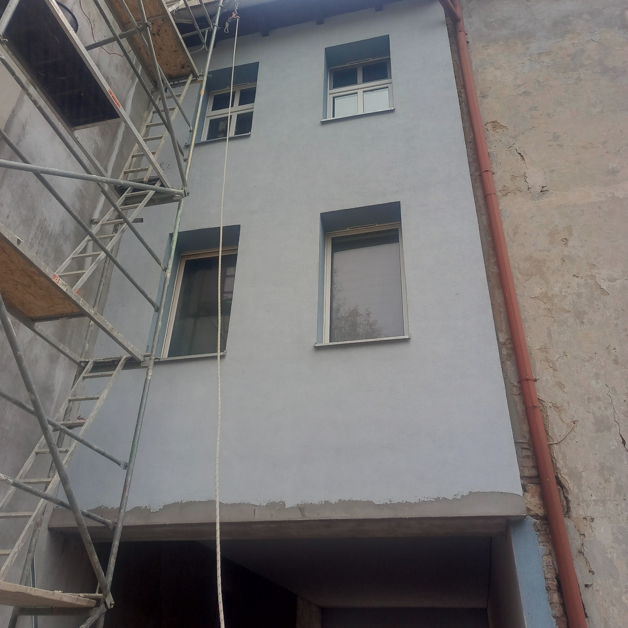 Świeżo pomalowana, błękitna elewacja budynku z oknami, widoczna z dołu. Fragment rusztowania po lewej stronie. Po prawej stronie widoczny fragment starej elewacji.