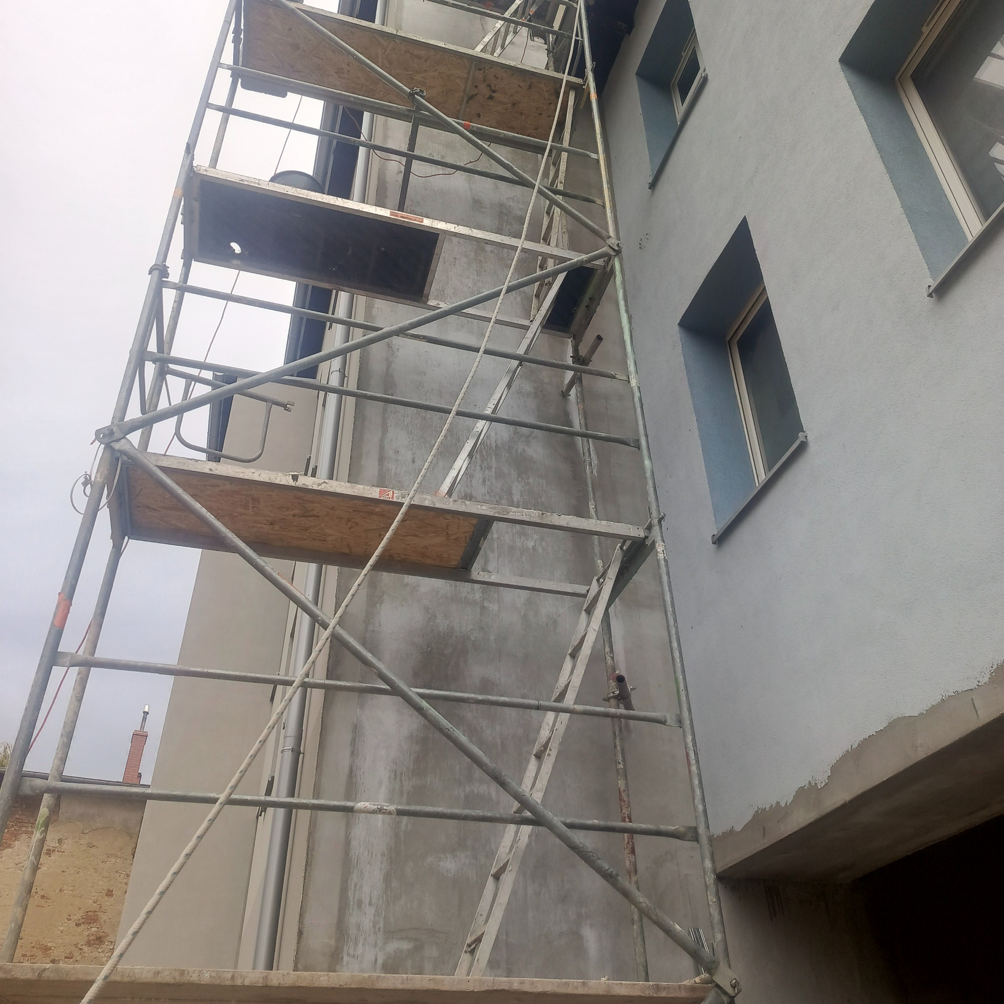 Elewacja budynku w trakcie prac wykończeniowych, widoczne rusztowanie i fragmenty otynkowanej ściany z oknami. Prace na wysokości.